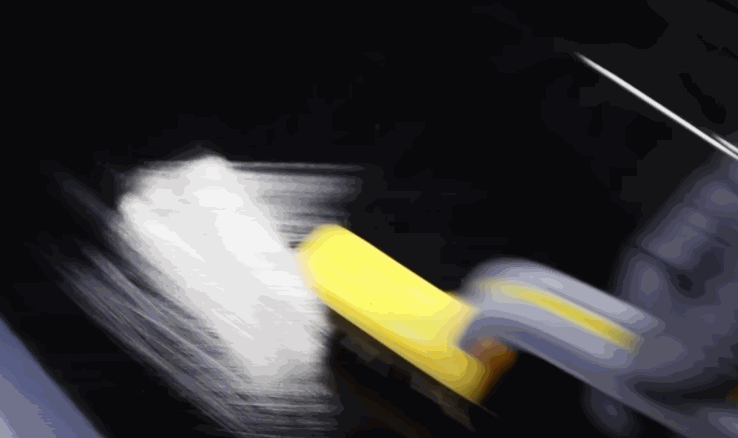 A hand with a yellow tool smooths paint protection film onto a glossy black car hood, blurring motion shows active squeegeeing for a bubble-free finish.