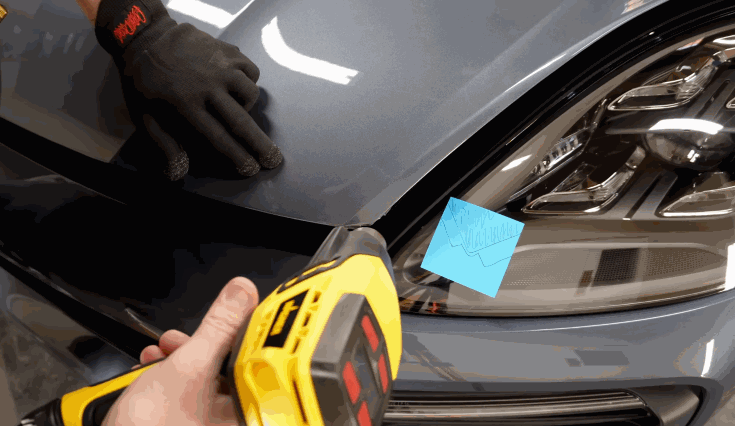 Installer uses a heat gun while smoothing paint protection film over the hood and headlight area of a grey car during professional installation.