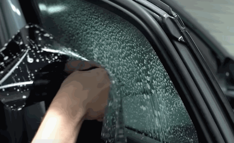Close up of hands pressing window tint onto a car window covered in water droplets, helping the film adhere evenly. The tint is being carefully positioned to ensure a clean and seamless fit.