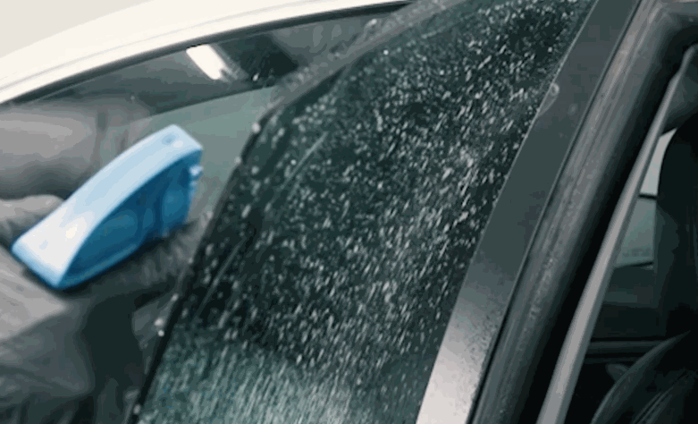 A blue squeegee rests on a wet Tesla window during window tint installation. The close up shows the film and moisture along the glass before final smoothing.