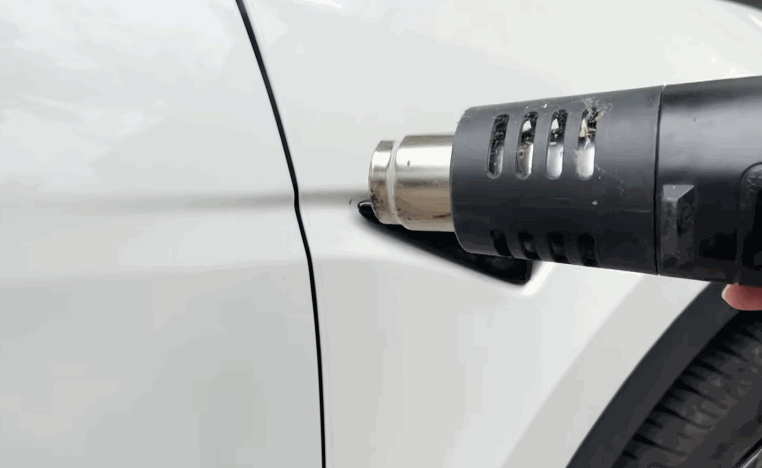 Heat gun used to shape paint protection film around a white vehicle door handle and body panel edge.