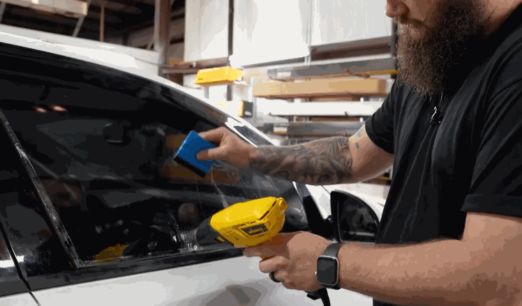 A person spraying solution onto a car window and using a squeegee to apply ceramic window tint, smoothing out the film.