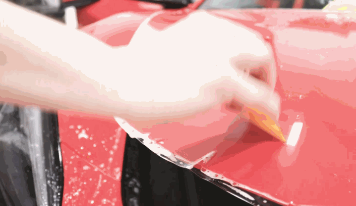 A person using a squeegee to install paint protection film on the hood of a red car.