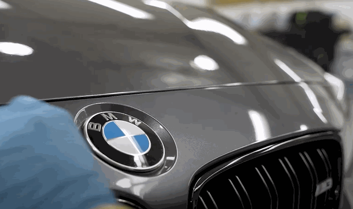 Close-up of a gloved hand applying ceramic coating to the hood of a silver BMW, focusing on the car's emblem and glossy surface reflection.