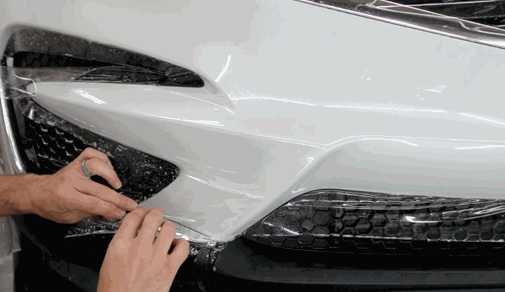 Close up of hands stretching and positioning paint protection film across the front bumper and grille of a white vehicle during installation.