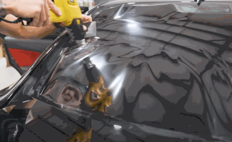 Technician uses a heat gun to apply window tint film smoothly across a car’s glossy black surface, removing wrinkles and air bubbles. The reflective finish shows careful precision during the tint installation process.