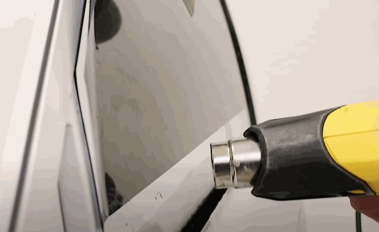 Technician uses a heat gun to apply window tint film smoothly across a car’s glossy black surface, removing wrinkles and air bubbles. The reflective finish shows careful precision during the tint installation process.