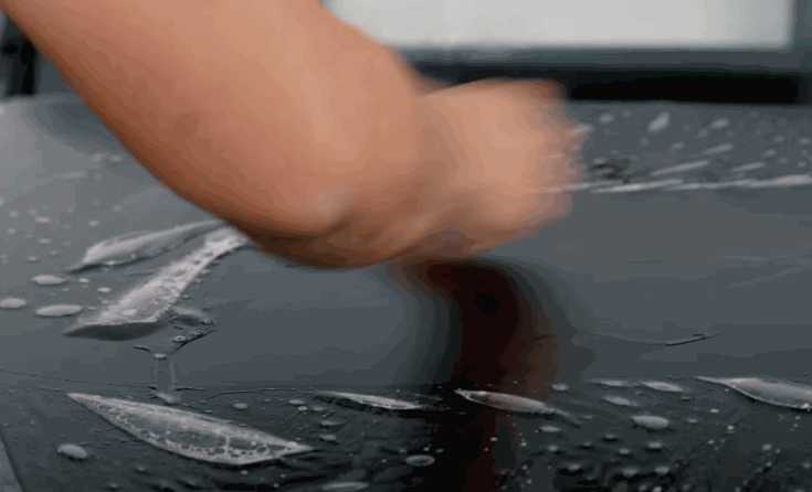 A technician smooths wet paint protection film across the hood of a dark vehicle during installation. Water droplets and film edges show the protective layer being positioned.