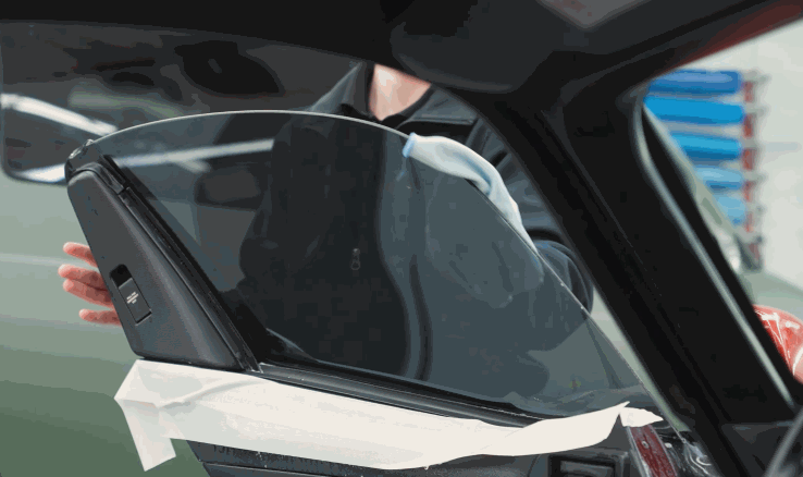 A technician positions window tint on an open car door window with tape along the lower edge. The side glass is being prepared for a clean installation.