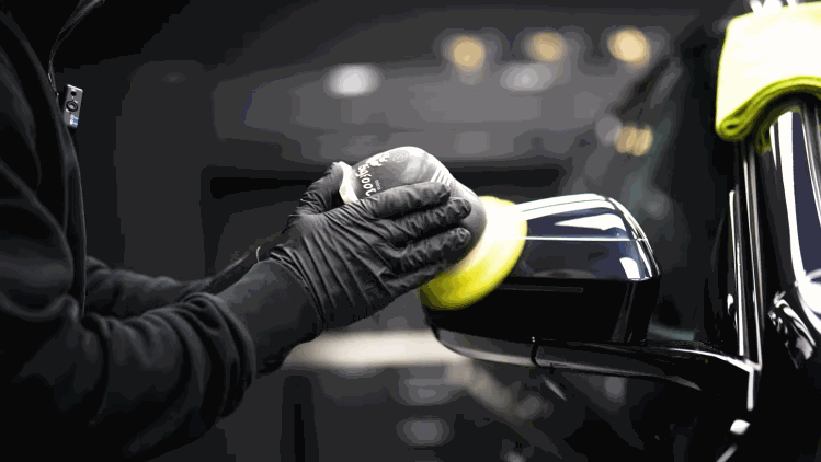 A technician hand polishes the side mirror of a black vehicle before ceramic coating. The close up shows careful detailing around the mirror and surrounding trim.