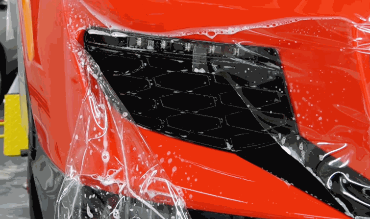 Close-up of paint protection film being applied to the front bumper vent area of a red vehicle, with moisture and bubbles visible during installation.