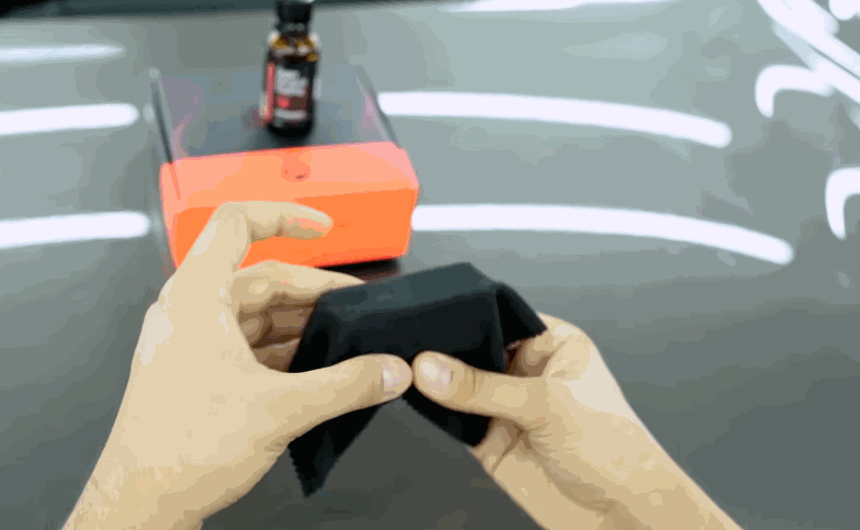 Hands prepare a small applicator cloth beside a coating bottle and foam block before applying ceramic coating to a vehicle.