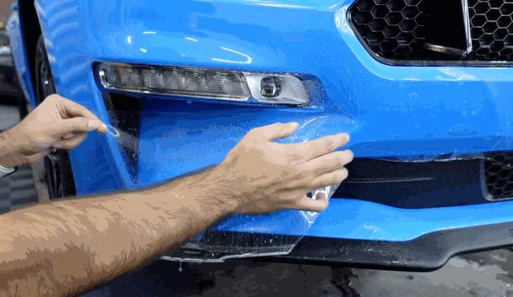 Hands stretch and position paint protection film over the front bumper of a bright blue car.