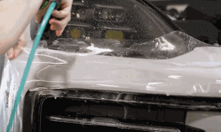 Technician stretching and aligning paint protection film over a car’s headlight and front panel.