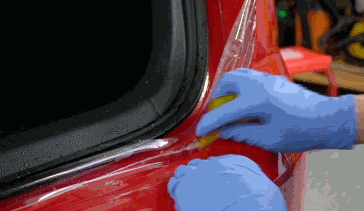A person in blue gloves uses a yellow squeegee to smooth paint protection film onto the rear panel of a red car.