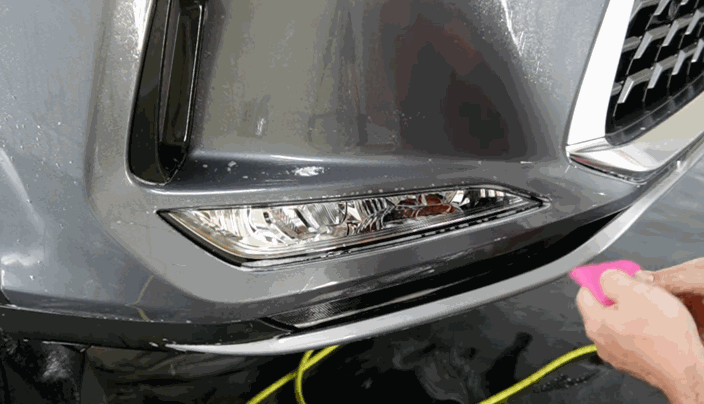 Technician installing paint protection film on a vehicle’s front bumper and headlight area using a pink squeegee.