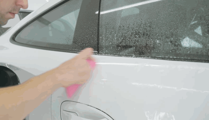 Technician applying paint protection film on a white car door with a pink squeegee, smoothing the surface to remove bubbles.