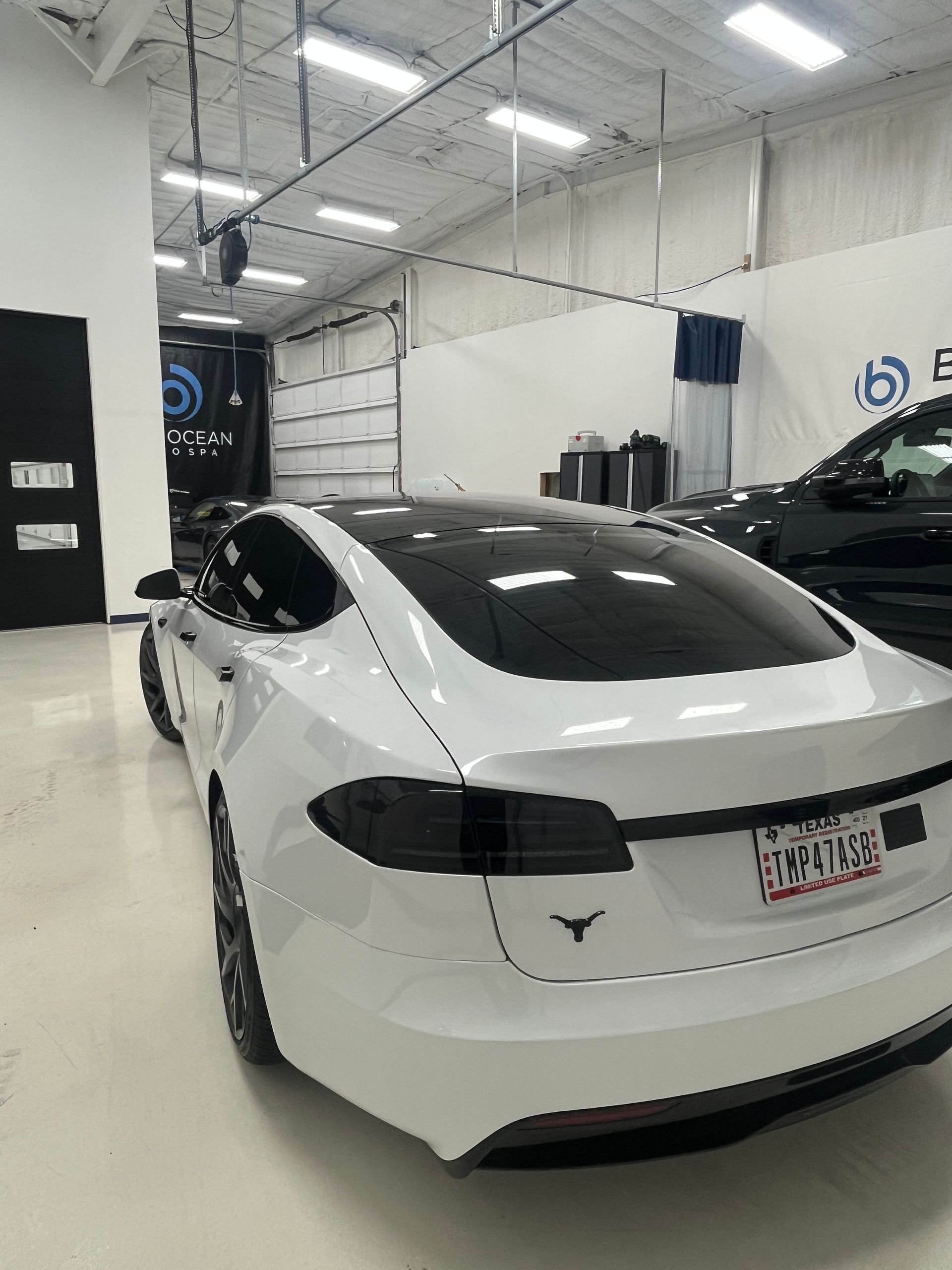 White Tesla in a bright auto shop garage, rear view with black-tinted windows and another car nearby