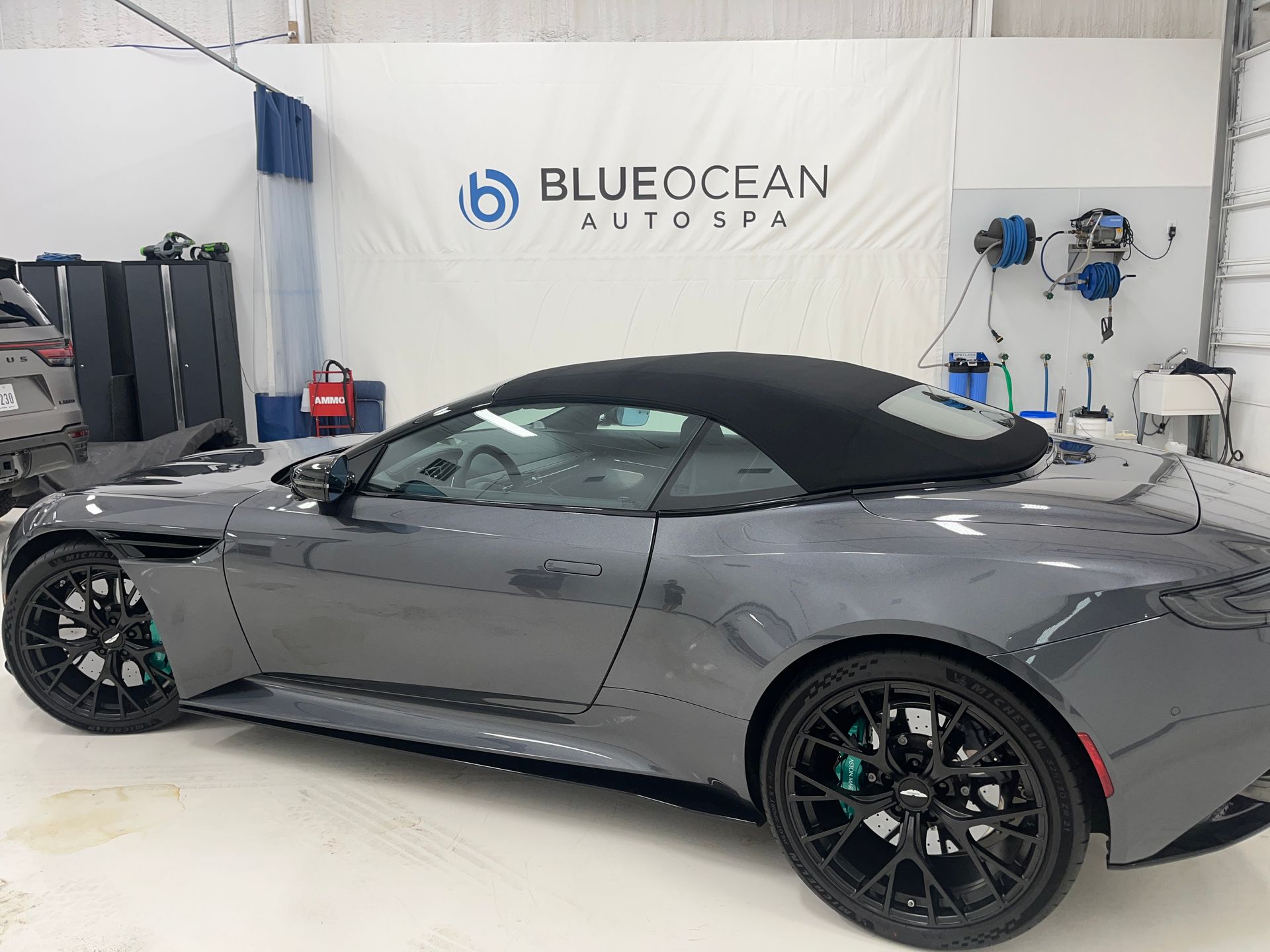 Gray Aston Martin convertible at a car wash with black top, black wheels, and green brake calipers.