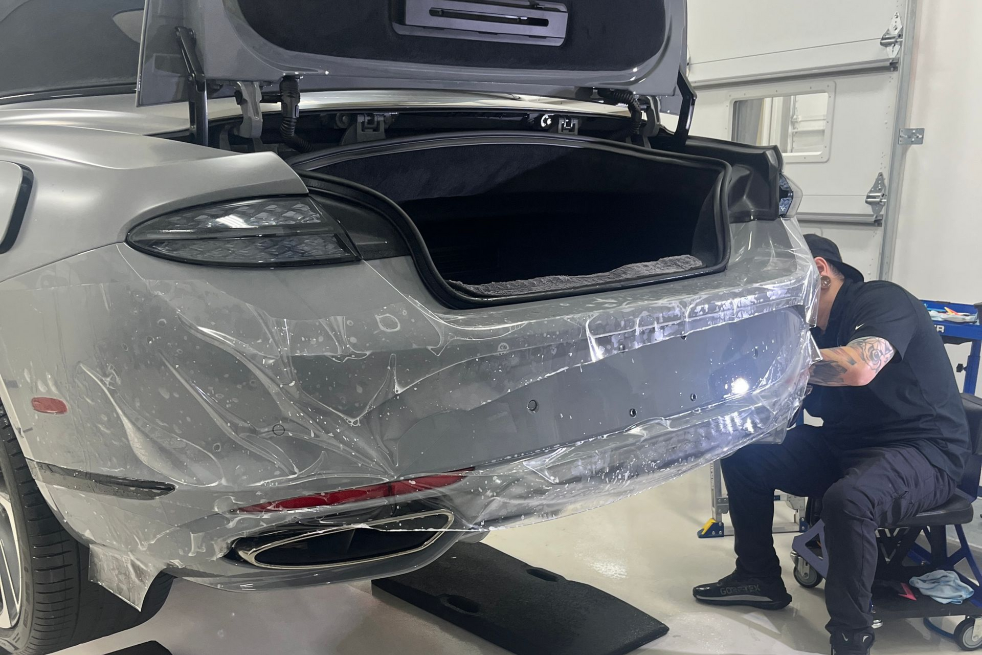 A man applies protective film to a black car's hood, using a spray bottle in a shop.