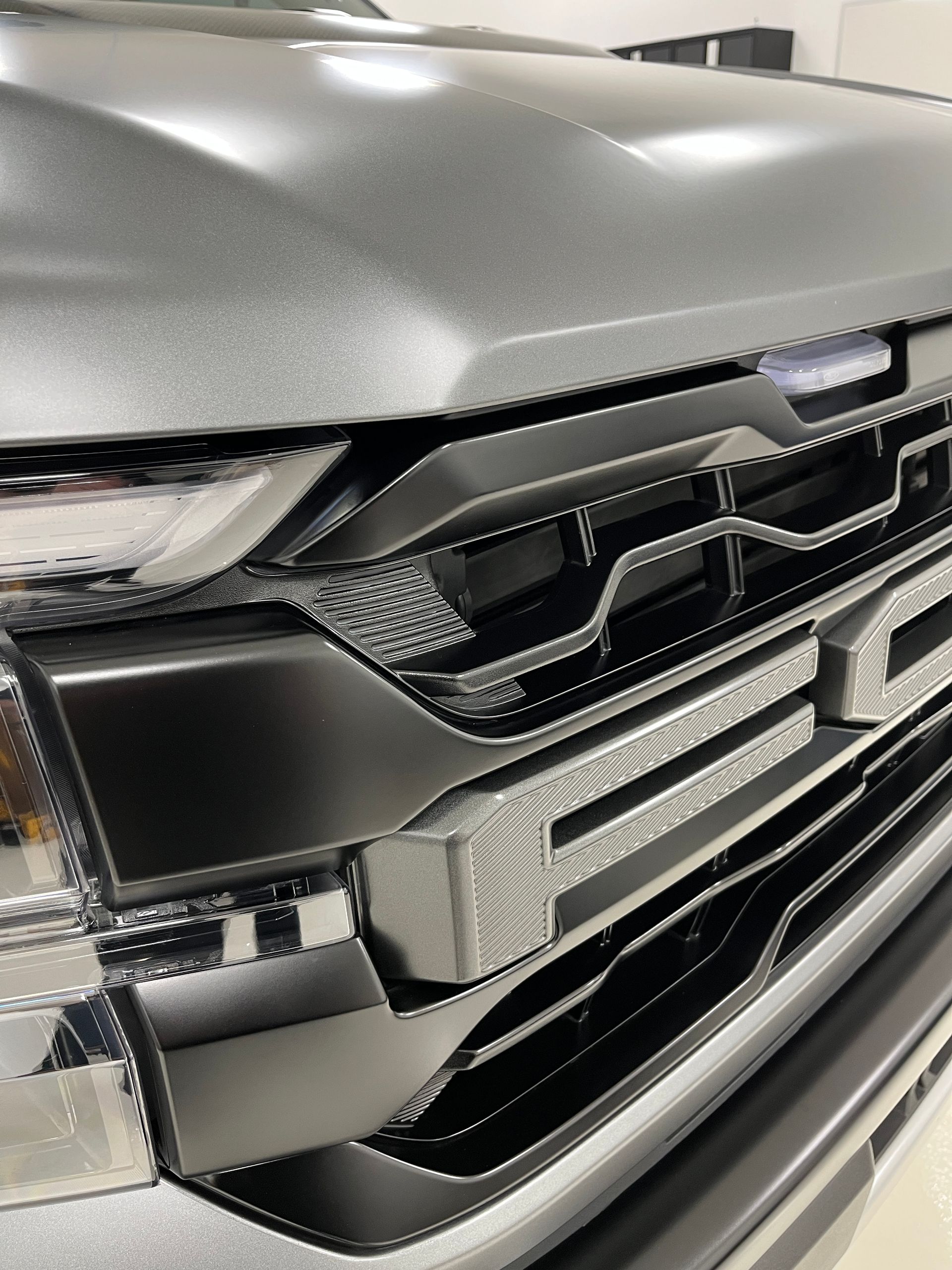 Close-up of a gray truck's front grill, showing a textured dark grill with a chrome emblem and details.
