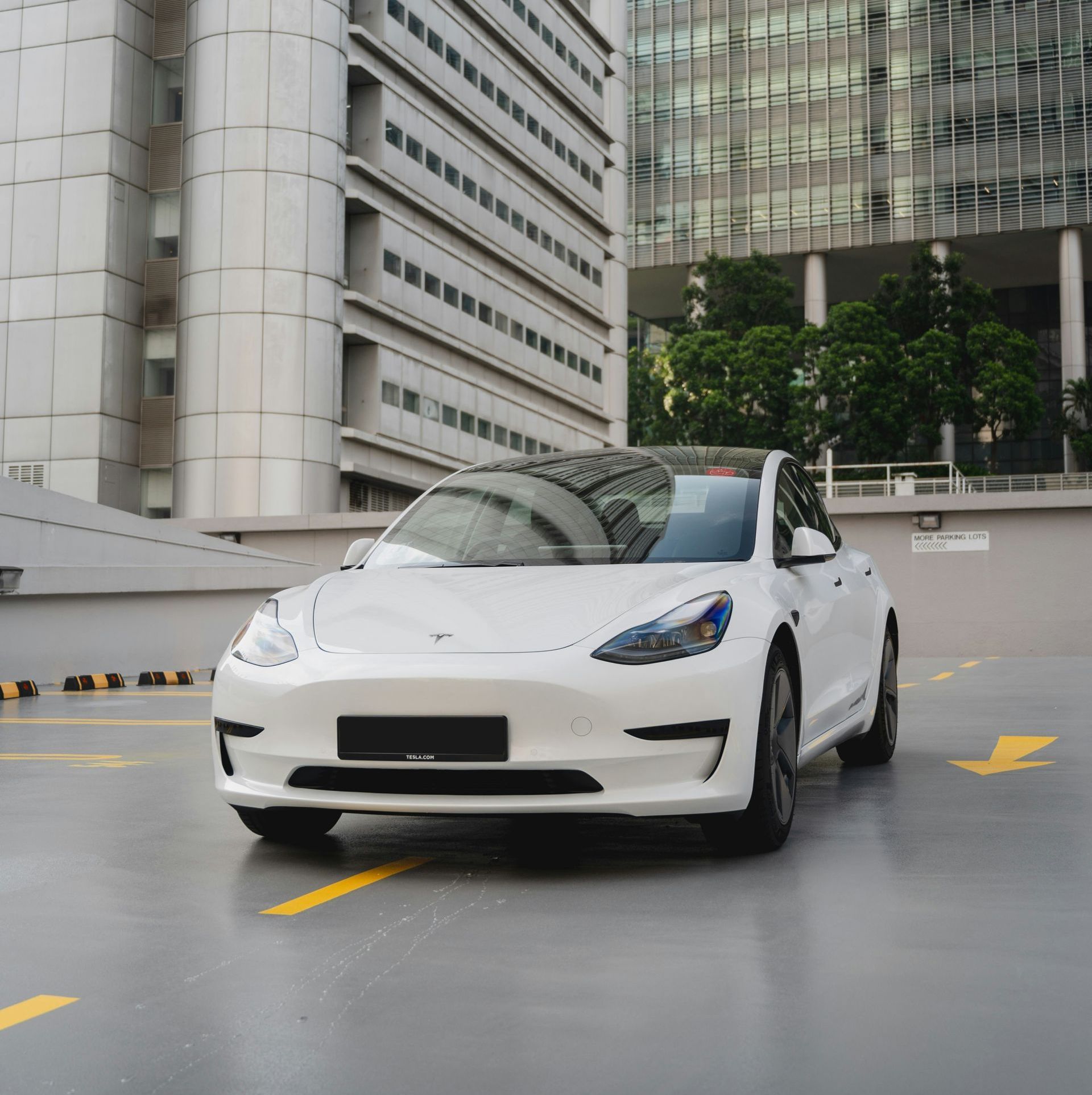 A white tesla model 3 is parked in a parking lot in front of a tall building.
