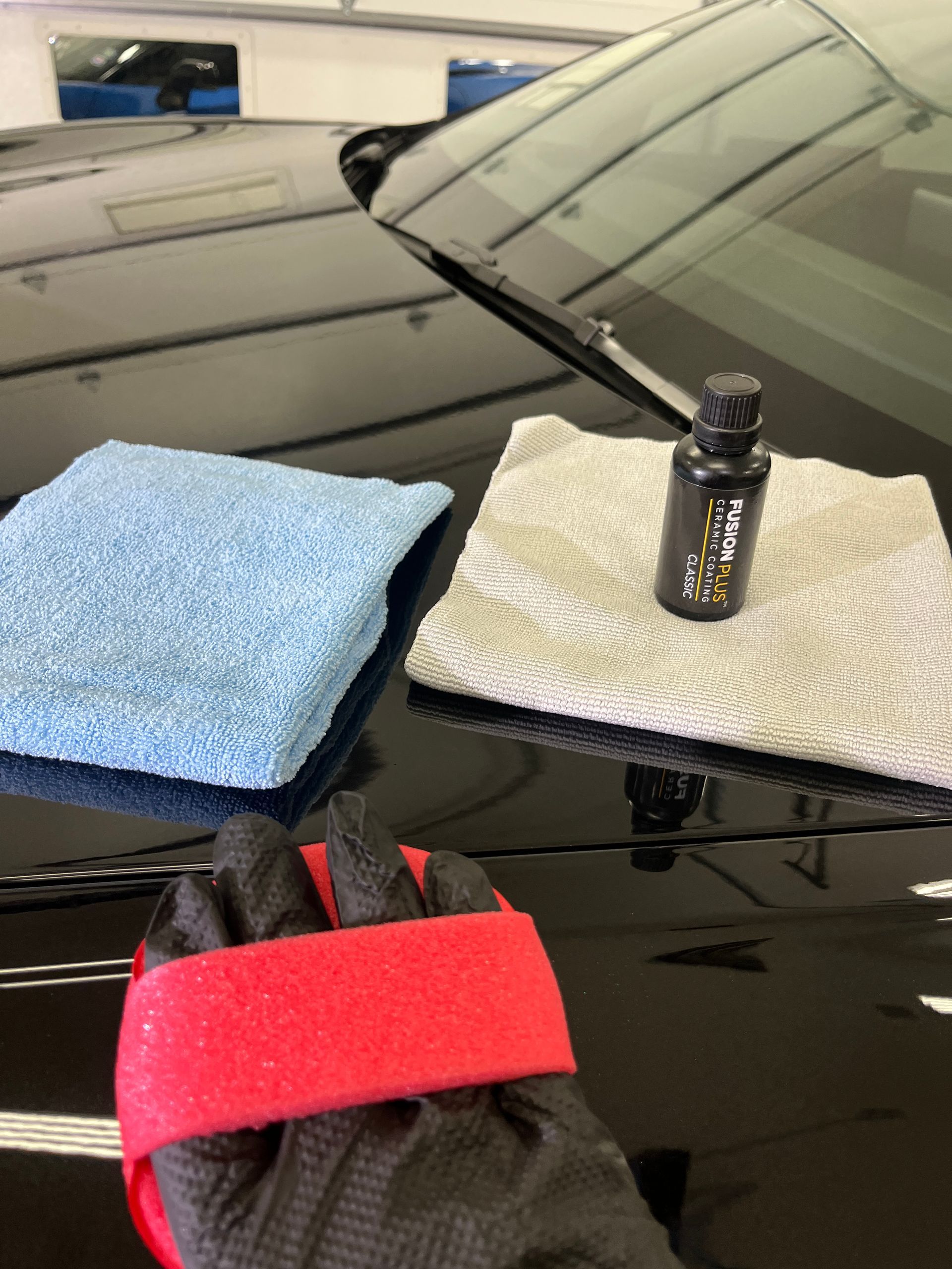 A gloved hand with a foam applicator pad is wiping on ceramic coating from the bottle in the picture.  Once the ceramic cross-links to the paint the pictured microfiber towels are used to level and wipe away the excess material.