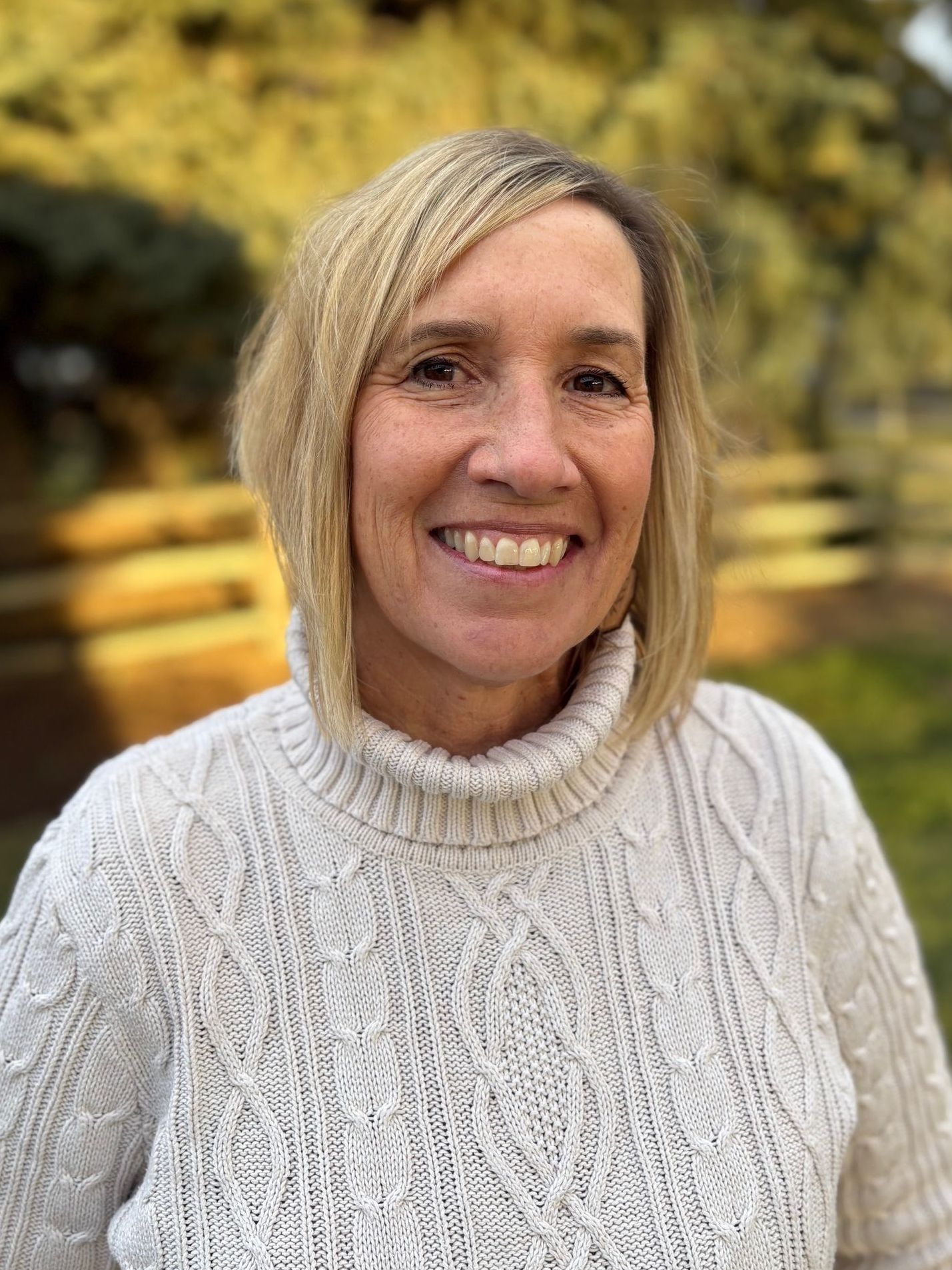 A woman in a white sweater is smiling in front of a tree.
