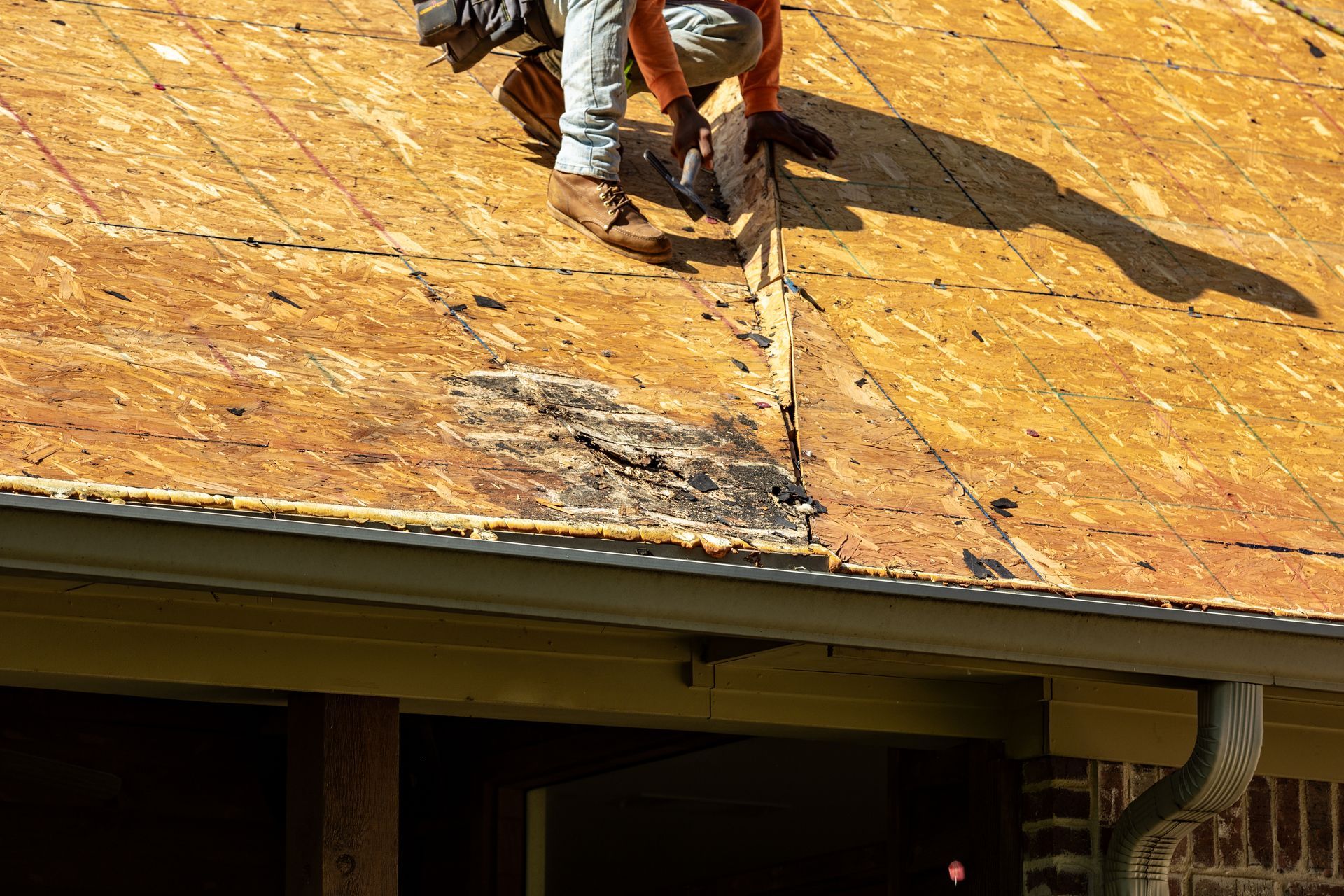 A roofer inspecting exposed decking with visible water damage and rot above the gutter