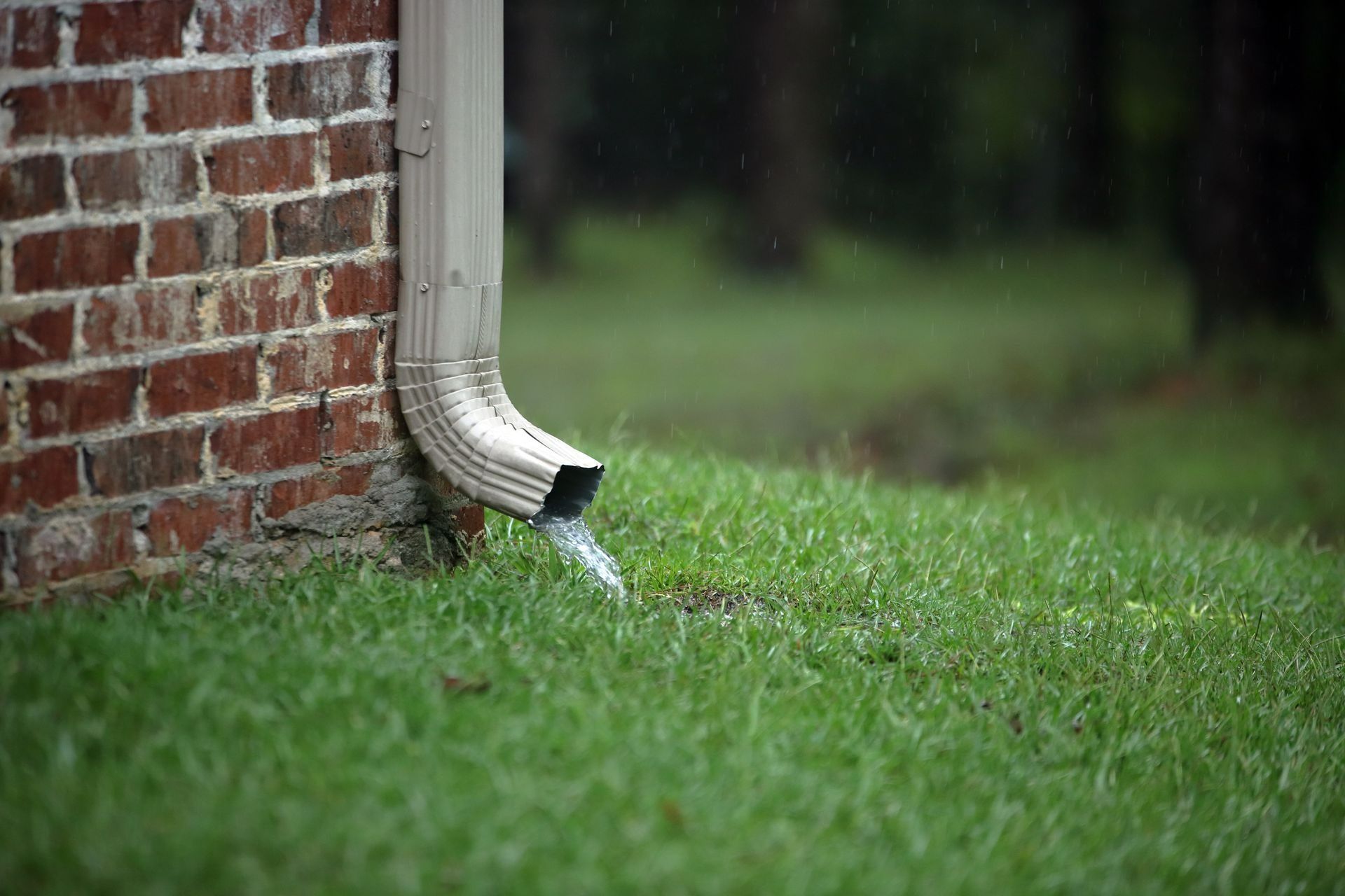 White downspout on a brick wall drains rainwater into a grassy yard.