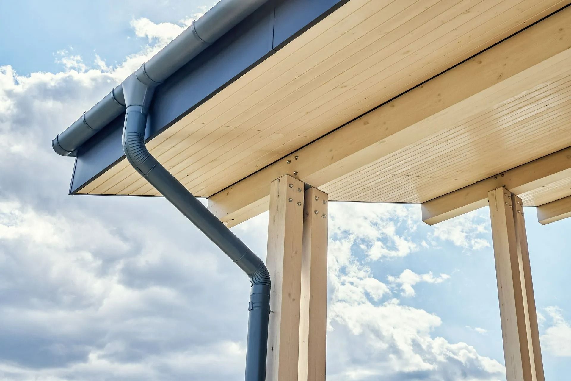 Close-up of a house roof gutter and downspout against a blue sky with clouds