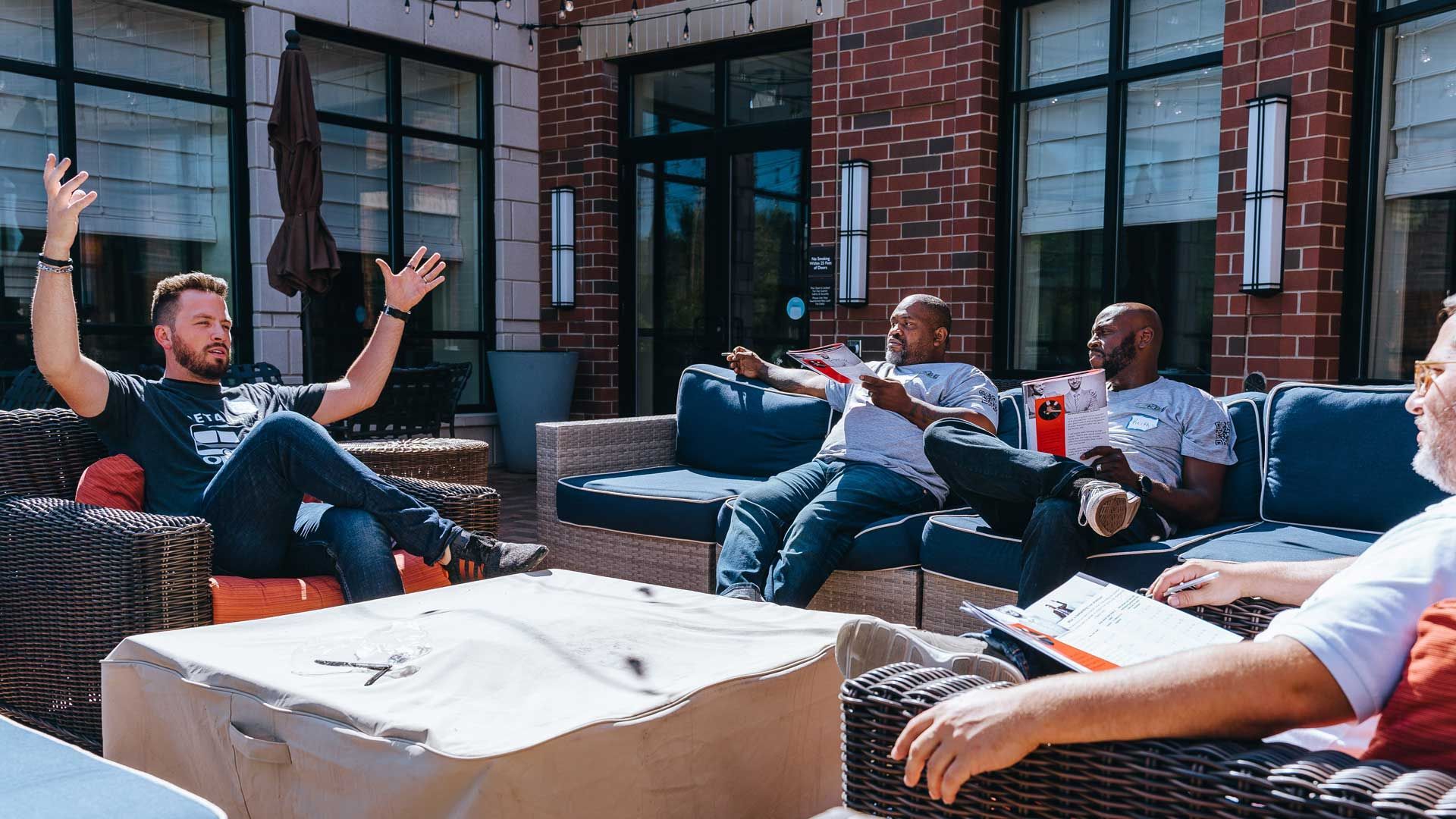 Four people seated outdoors on patio furniture, gesturing and talking near a brick wall.