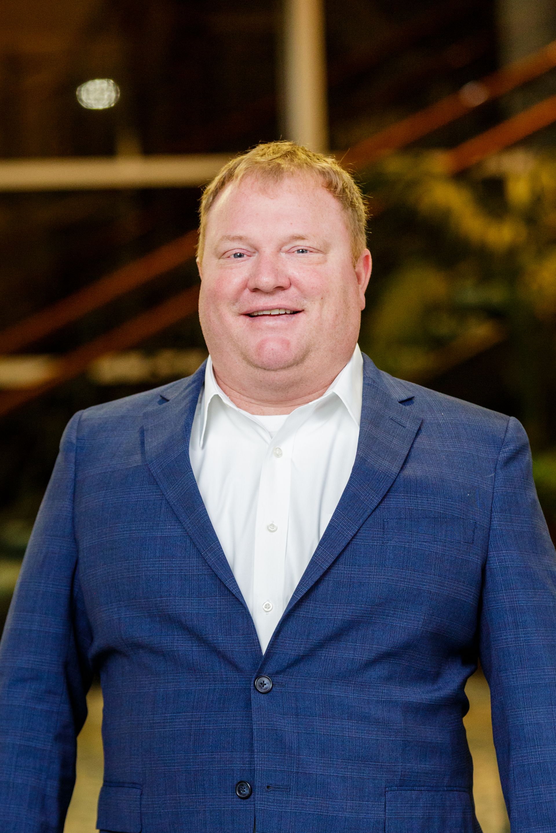Smiling man in a blue blazer and white shirt standing indoors against a blurred background