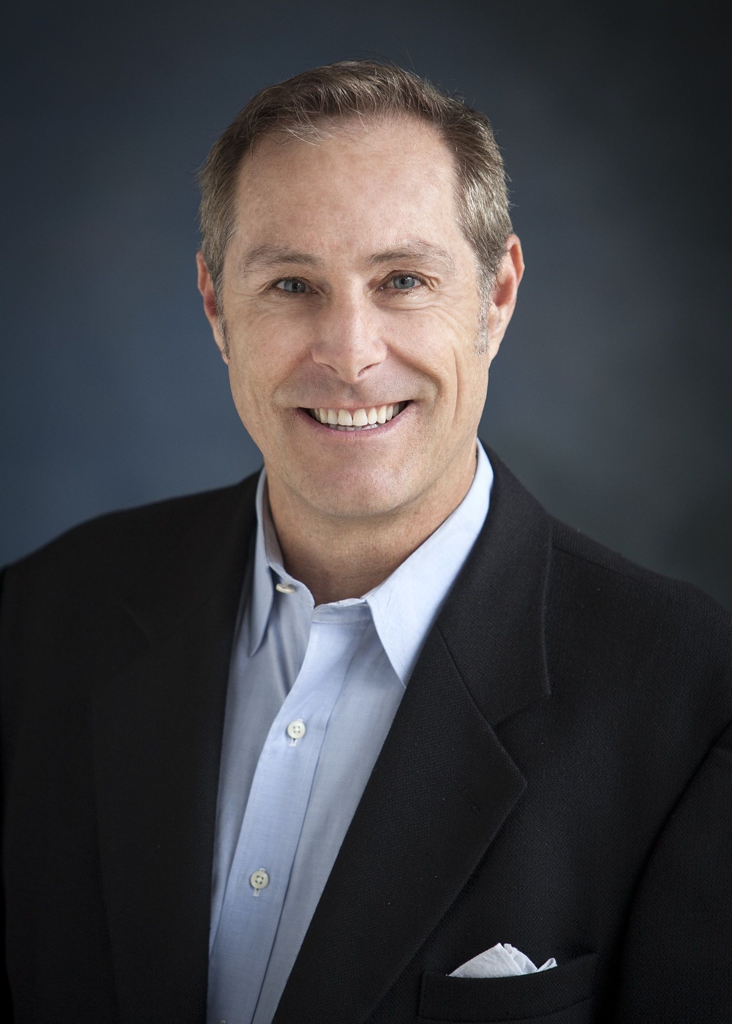 Smiling man in a dark suit and light blue shirt against a dark studio background