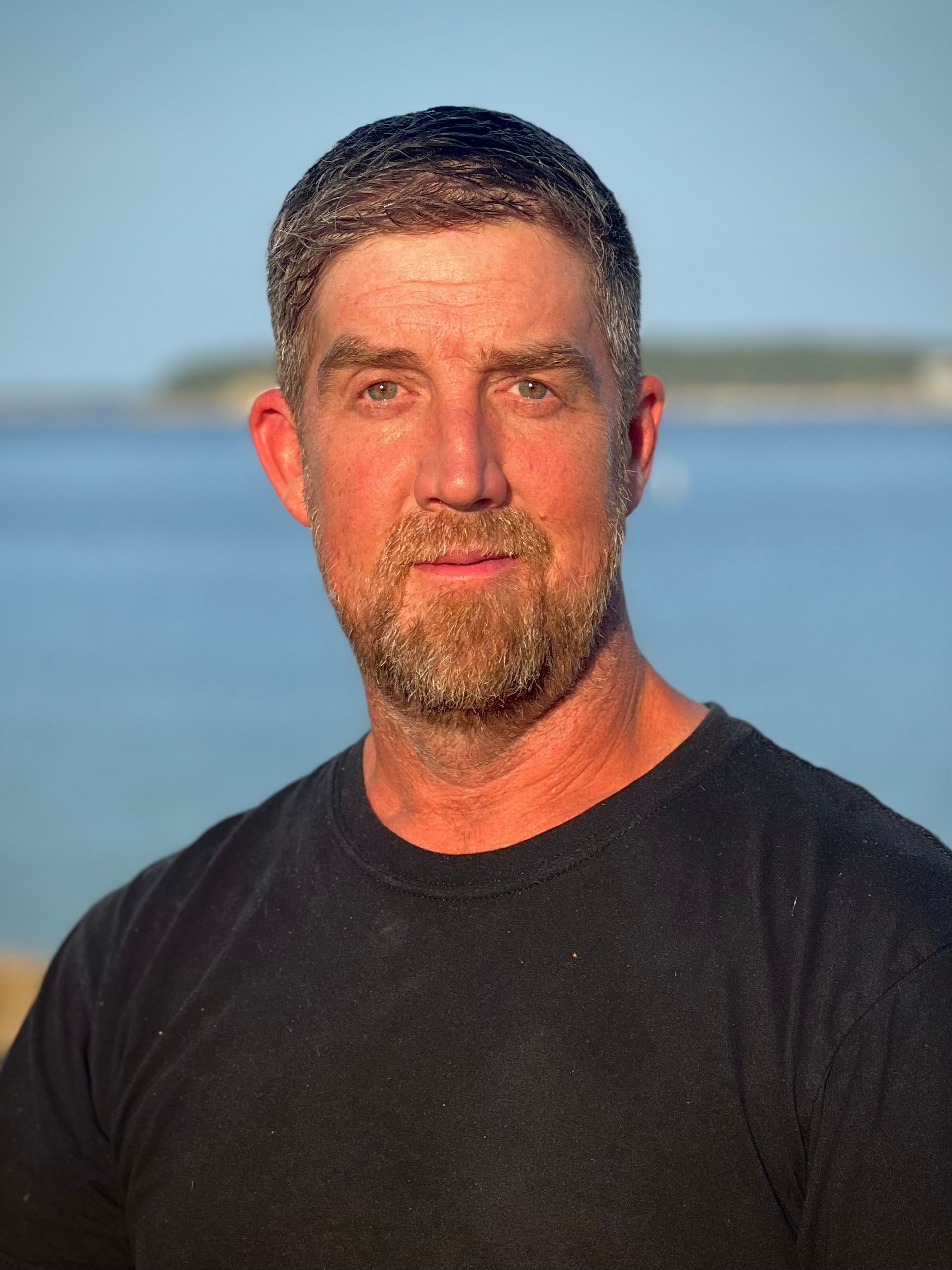 Man in a black shirt standing outdoors at the beach during sunset, with blue sky and ocean behind him