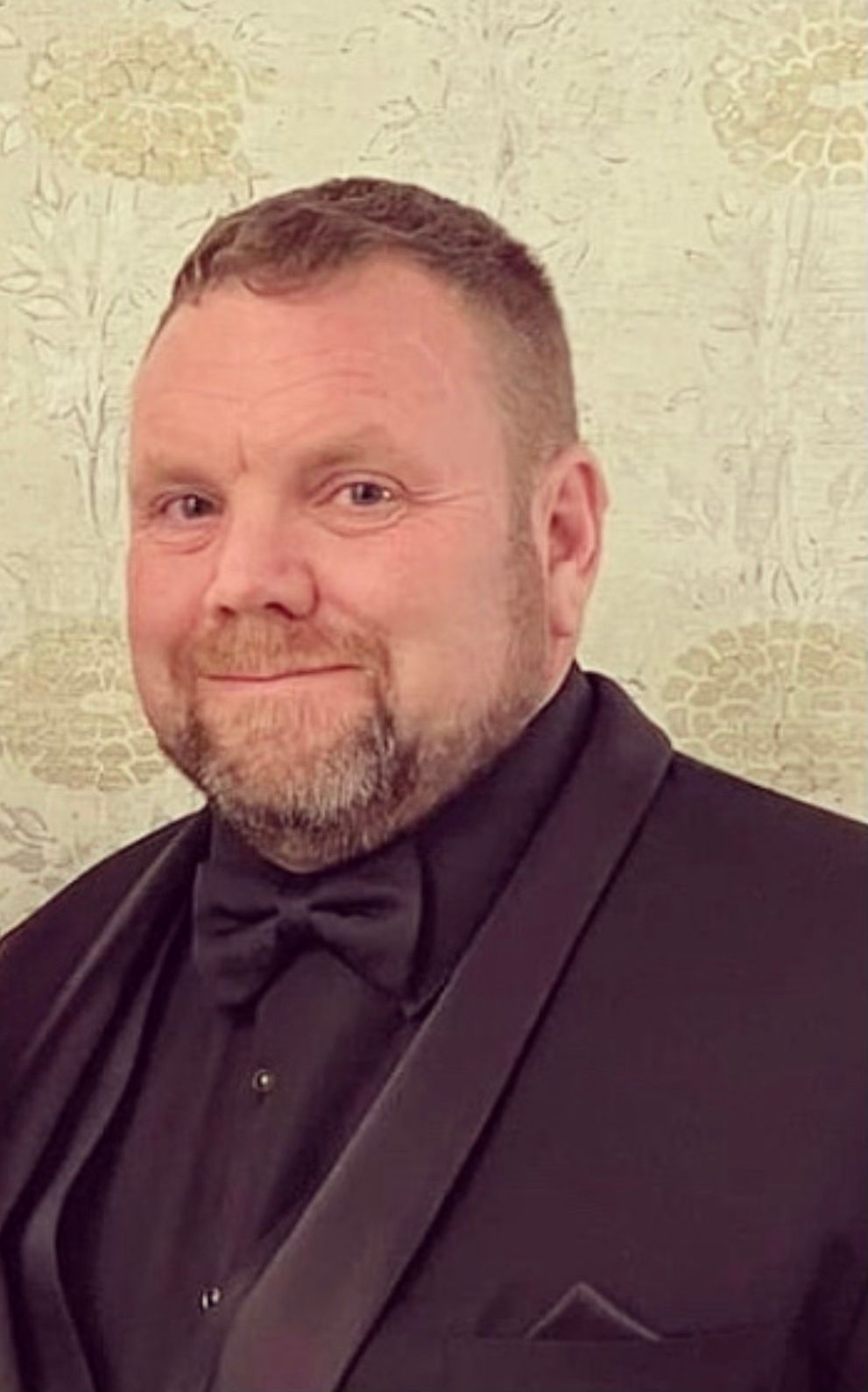 Man in a black suit and bow tie, smiling against a light textured wall.