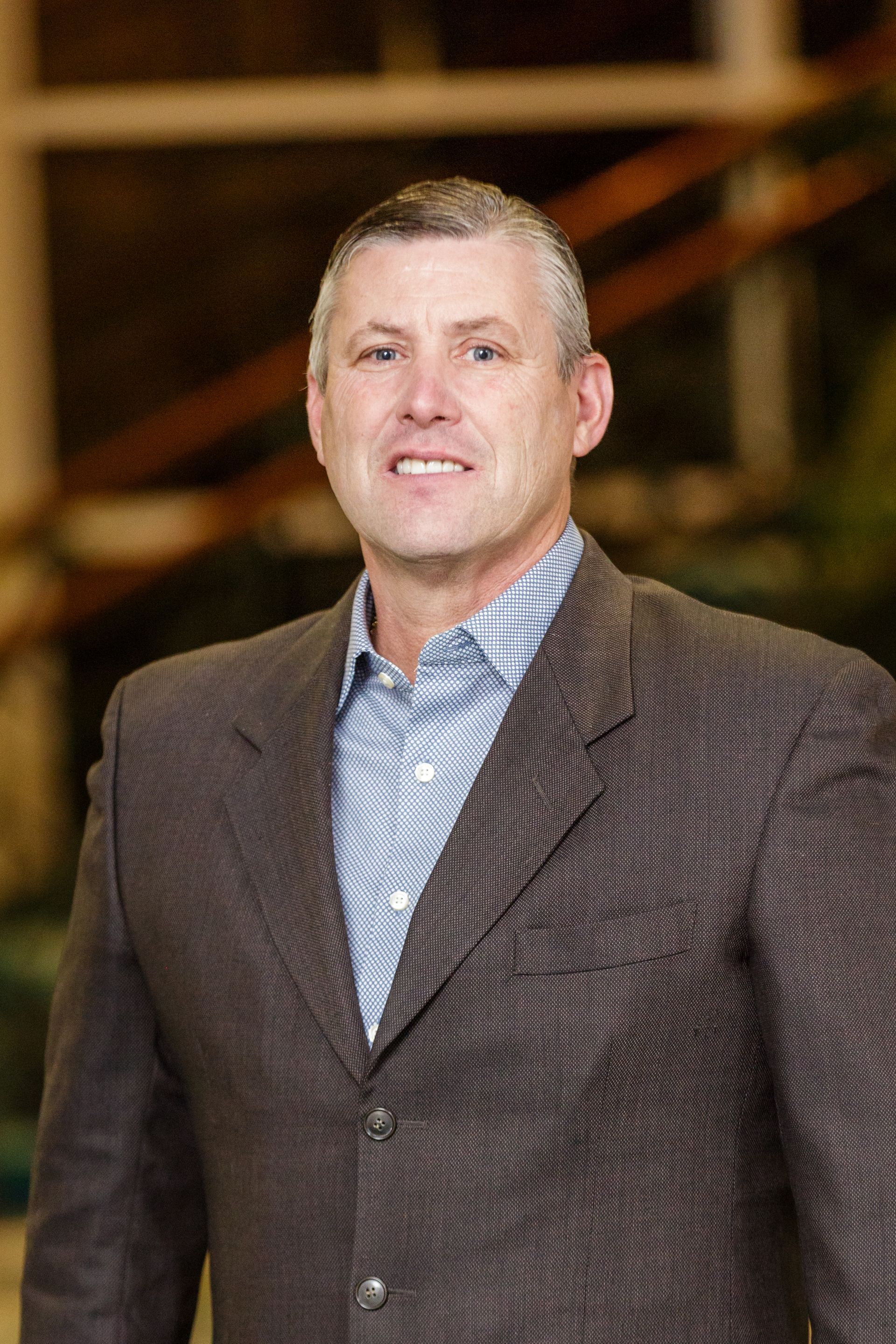 Man in a patterned blazer and blue shirt standing indoors, with a blurred staircase in the background