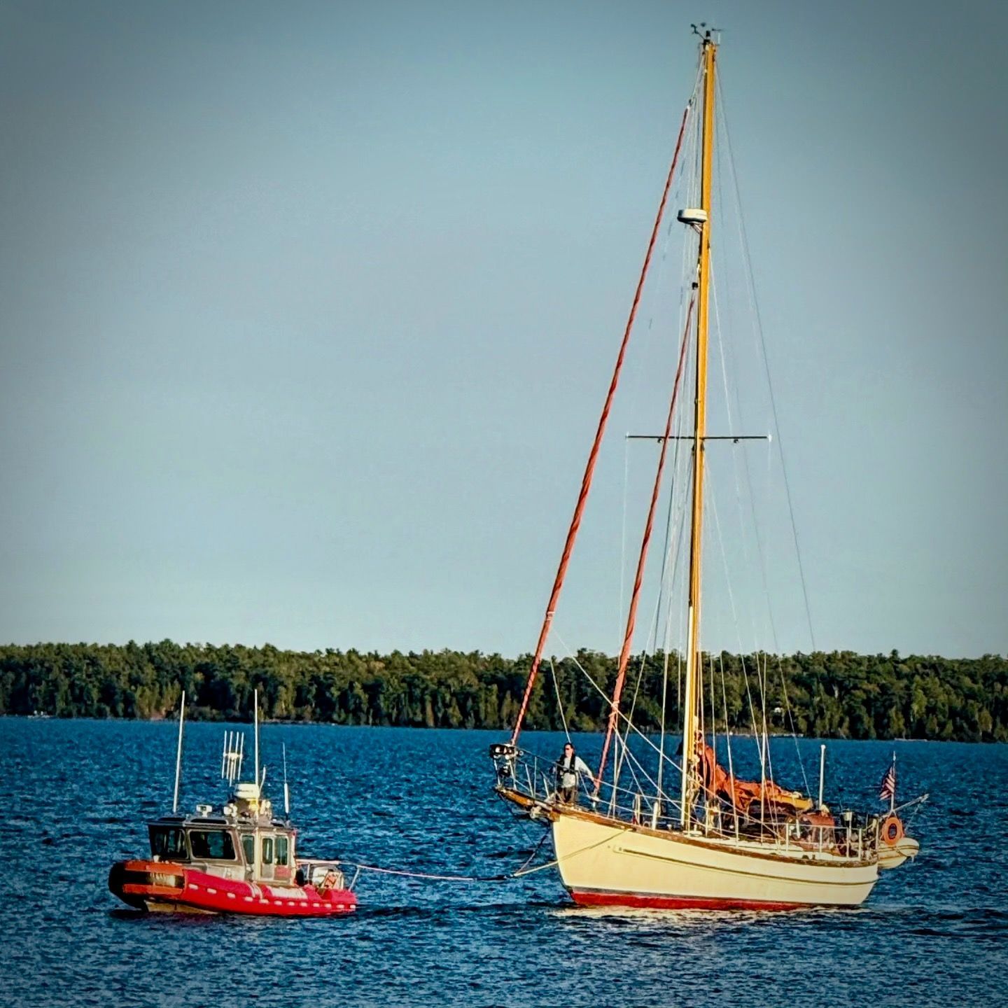 Red tugboat beside a sailboat on calm blue water under a clear sky