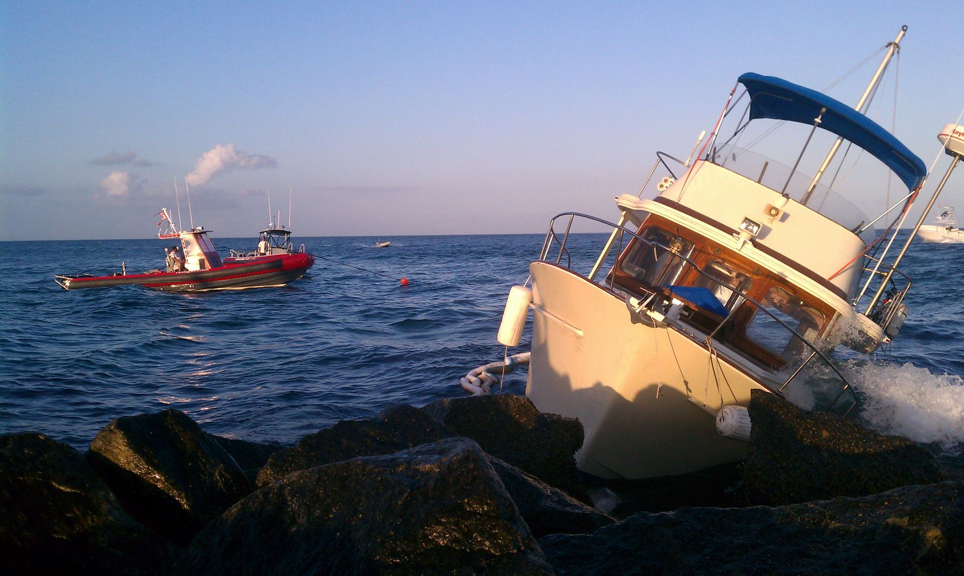 White boat listing on rocks in rough sea, with a red rescue boat nearby at sunset