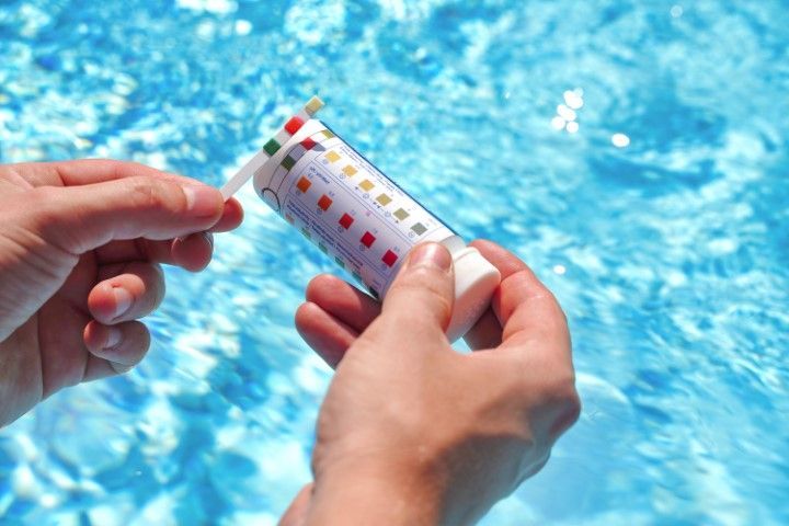 Hands holding a pool water test strip above blue swimming water