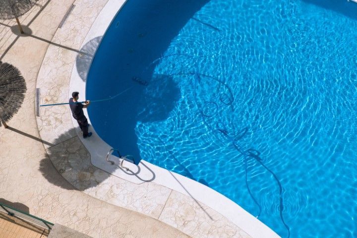 Person standing by a curved blue swimming pool, holding a hose on a tiled deck