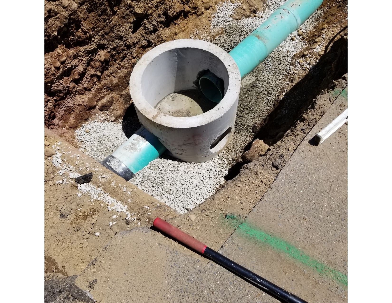 Concrete manhole and pipes in a construction site trench, surrounded by gravel and dirt.