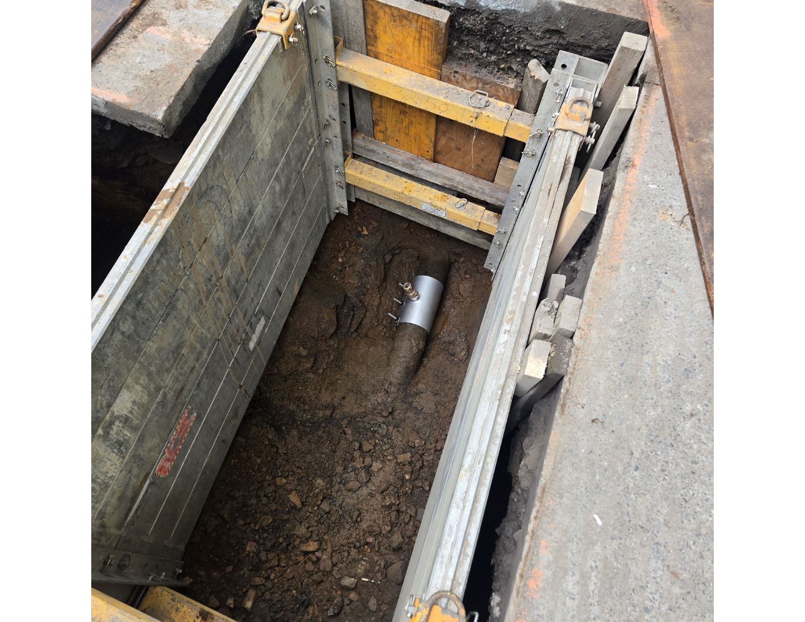 Trench box in excavation with a pipe visible. Grey metal, yellow supports, brown soil.