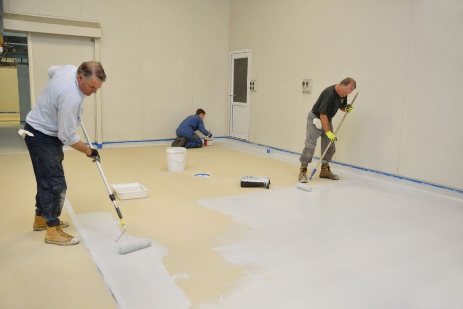 Workers painting a large empty room floor white with rollers and buckets