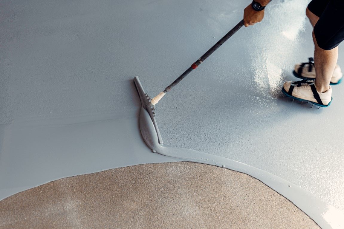 Person smoothing gray floor coating with a squeegee on a light concrete surface
