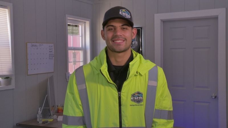 Smiling worker in a neon yellow safety jacket and black cap standing indoors near a door and calendar.