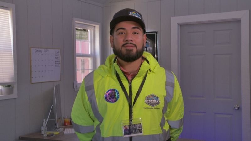 Person in a neon yellow safety jacket and cap standing indoors in a room with a door and calendar.
