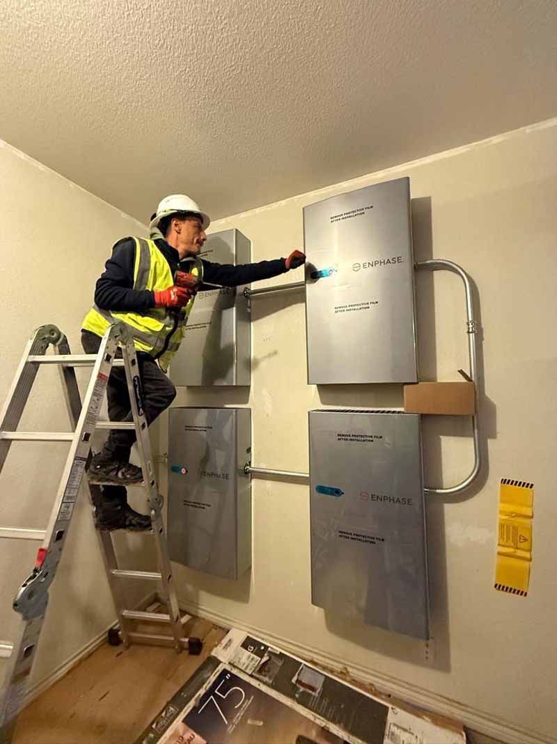 Worker in safety gear inspecting wall-mounted electrical panels from a ladder in a utility room