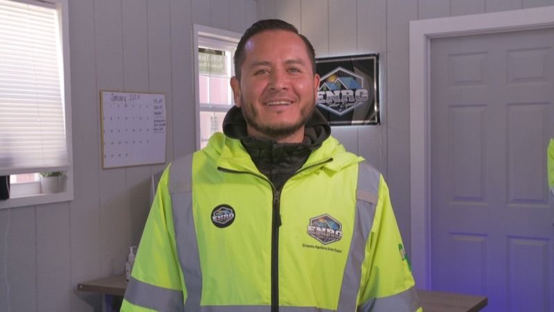 Smiling man in a neon yellow jacket standing indoors near a doorway and poster