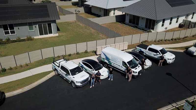 Several parked cars on a residential street with people standing beside them and one white van nearby